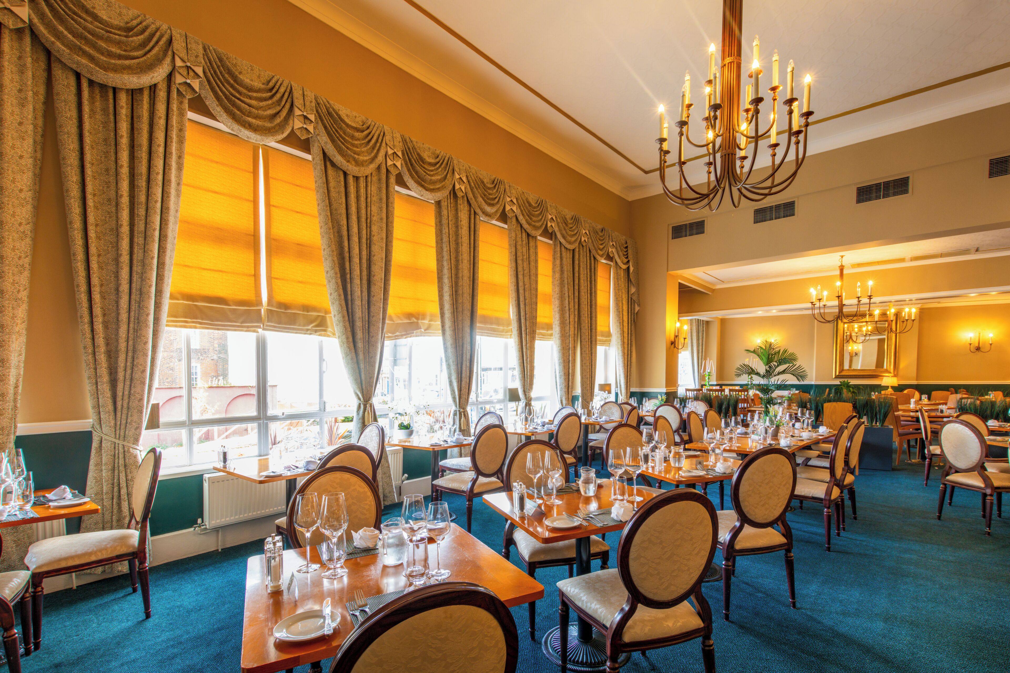 A hotel dining room with tables set for service, large windows with curtains, and a chandelier.