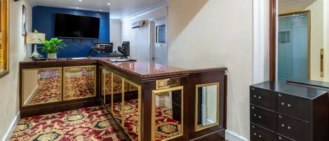 A hotel lobby with a reception desk, a large flat-screen TV, a mirror, a potted plant, and a dark wooden cabinet with drawers.