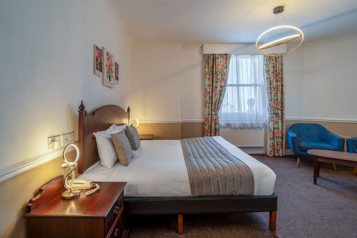 A hotel room with a bed, bedside table, lamp, chair, and curtains.