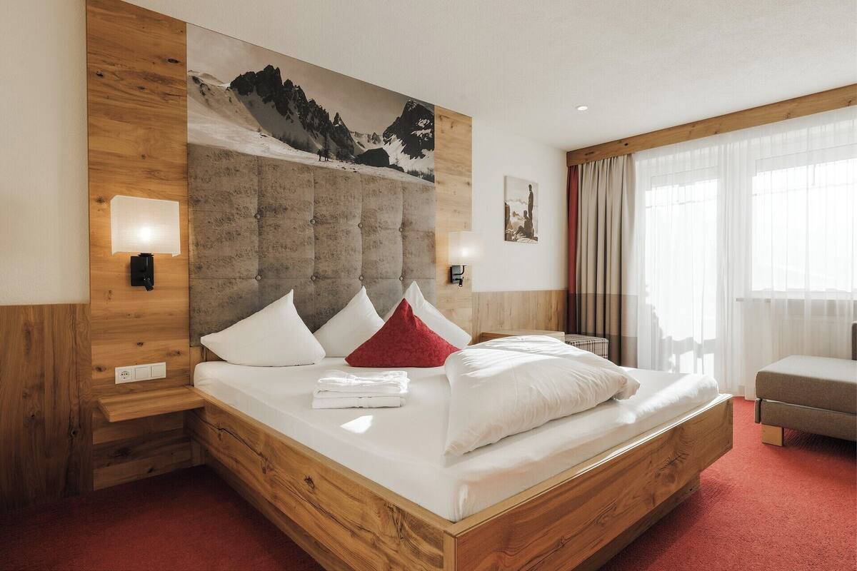 Ein Hotelzimmer mit einem Bettgestell aus Holz, einem großen Bergbild an der Wand, einem Nachttisch aus Holz und einem Fenster mit Vorhängen.