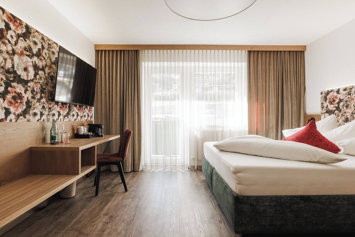 Ein Hotelzimmer mit Bett, Schreibtisch, Stuhl, einem an der Wand befestigten Fernseher und einem Fenster mit Vorhängen.