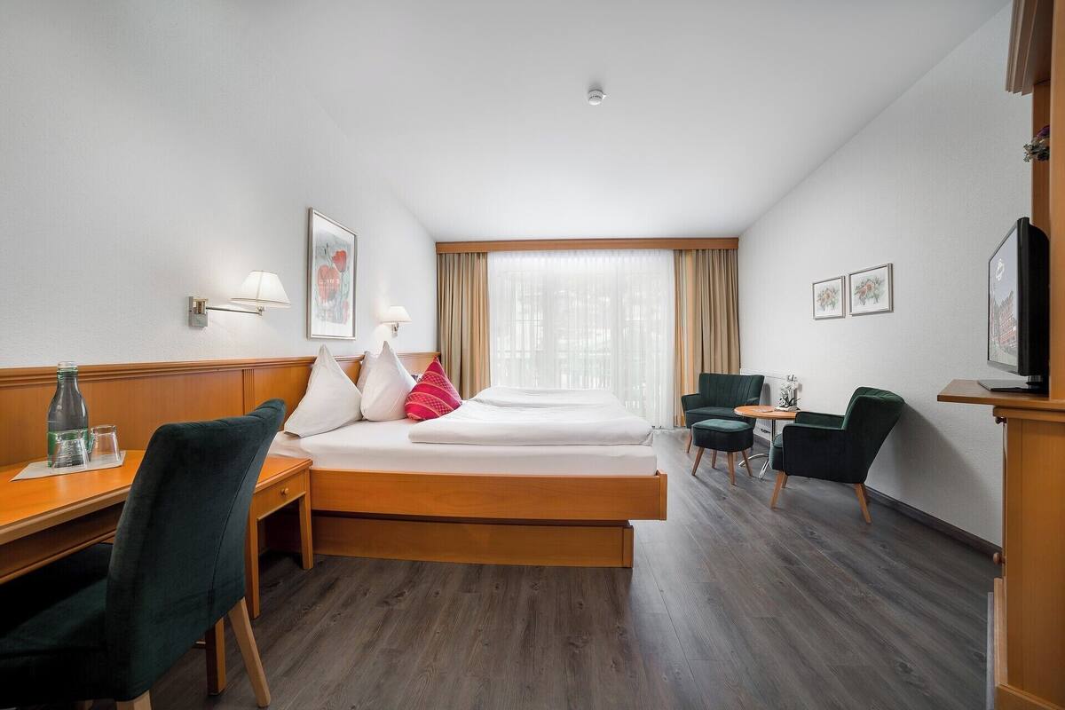 Ein Hotelzimmer mit einem Bett, zwei Sesseln, einem Schreibtisch, einem Fernseher und einem Fenster mit Vorhängen.