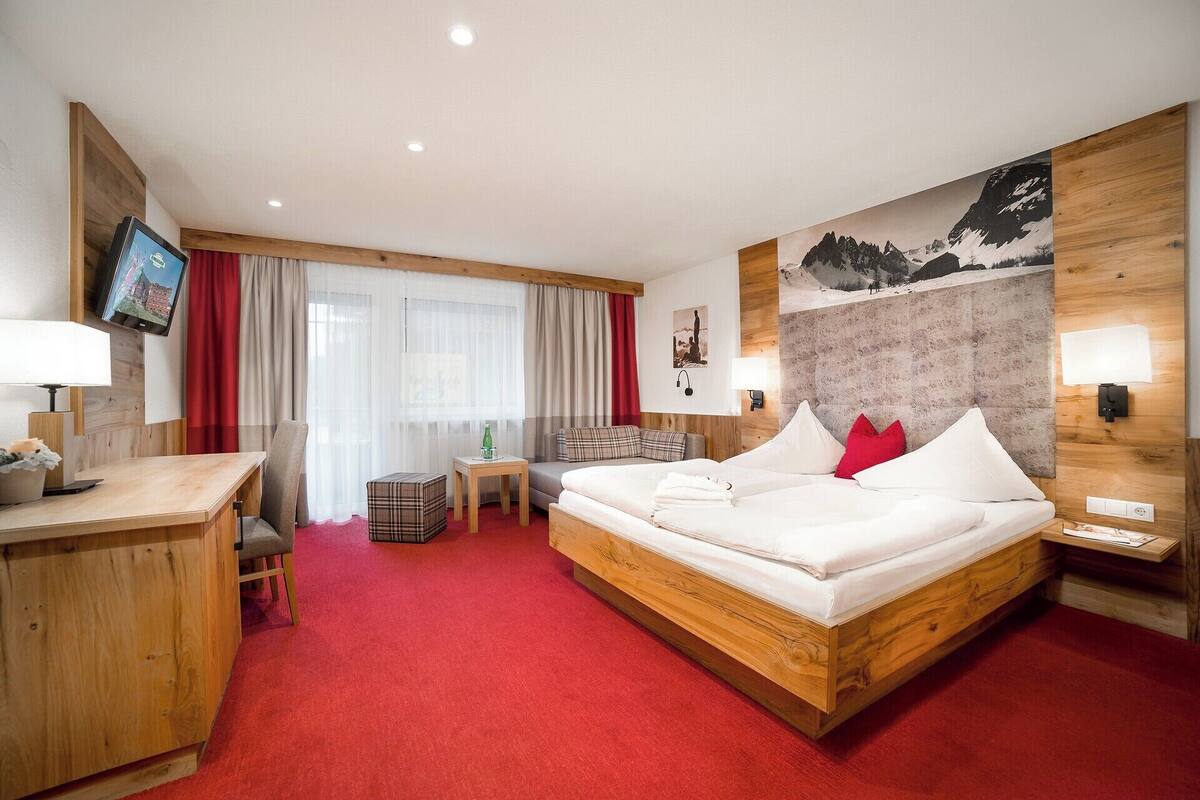 Ein Hotelzimmer mit einem hölzernen Bett, einem Schreibtisch, einem Fernseher und einem großen Fenster mit Vorhängen.