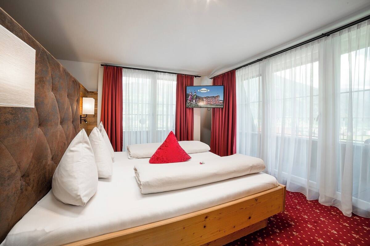Ein Hotelzimmer mit einem hölzernen Kopfteil, einem Bett mit weißen Leinen und einem roten Kissen, einem angebrachten Fernseher und großen Fenstern mit durchsichtigen Vorhängen.