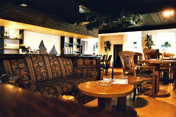 A cozy lounge area with a sofa, a round wooden table, and a bar area in the background. - Edelweiß (Goetzens)
