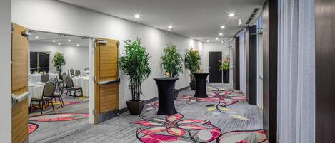 A hotel corridor with a floral carpet, potted plants, and a conference room in the background.