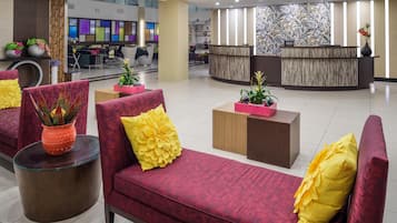 Eine moderne Lobby mit einer Rezeption, roten Sofas mit gelben Kissen und verschiedenen Kübelpflanzen.
