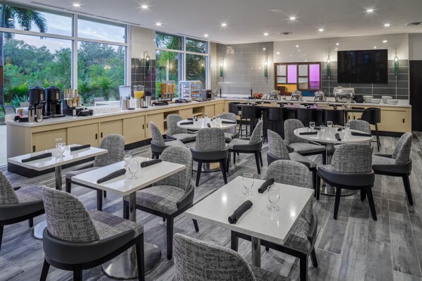 Un restaurante moderno con mesas y sillas redondas, una zona de bar y un ventanal grande con vistas a zonas verdes.