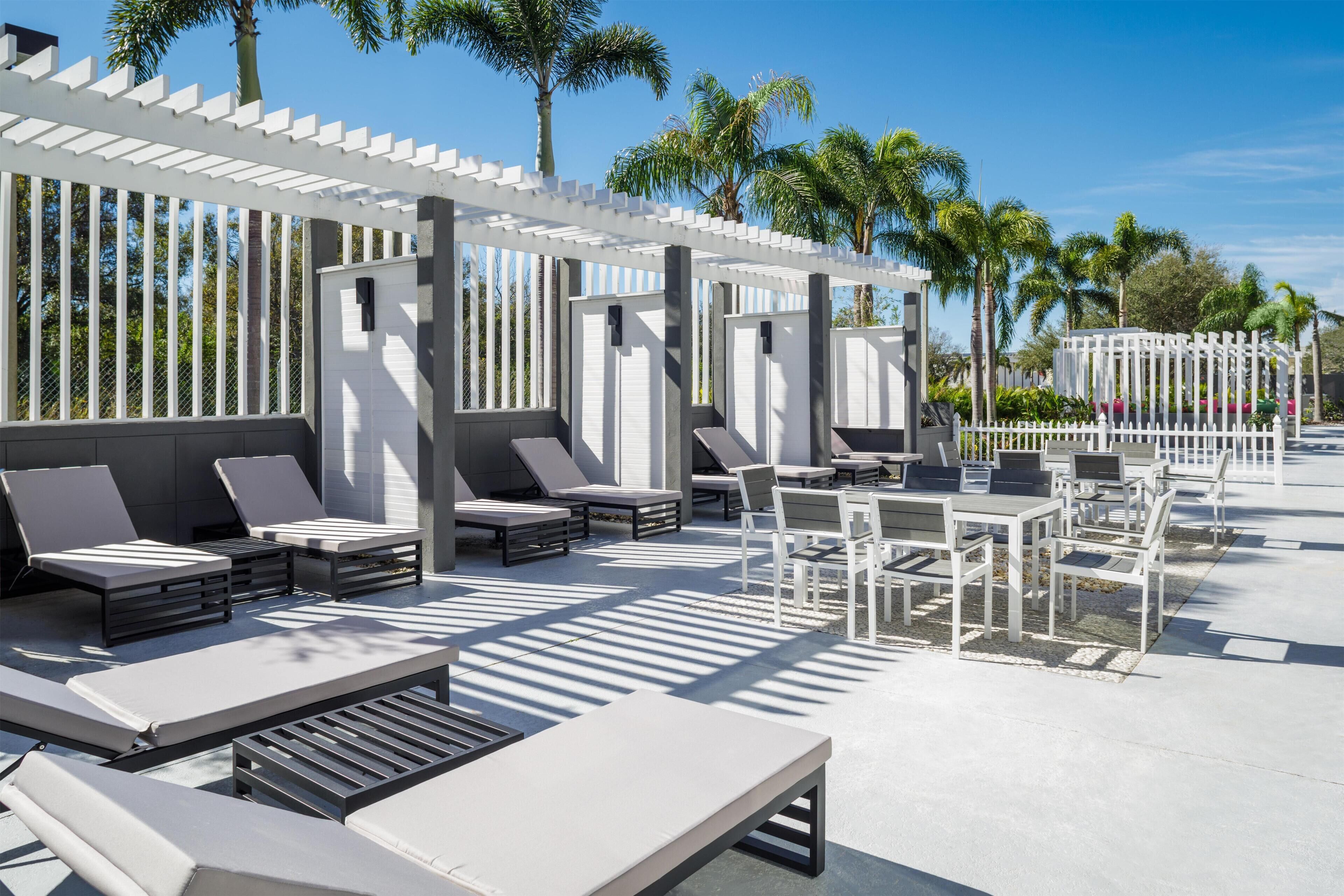 An outdoor seating area with white furniture, including lounge chairs and tables, under a pergola.