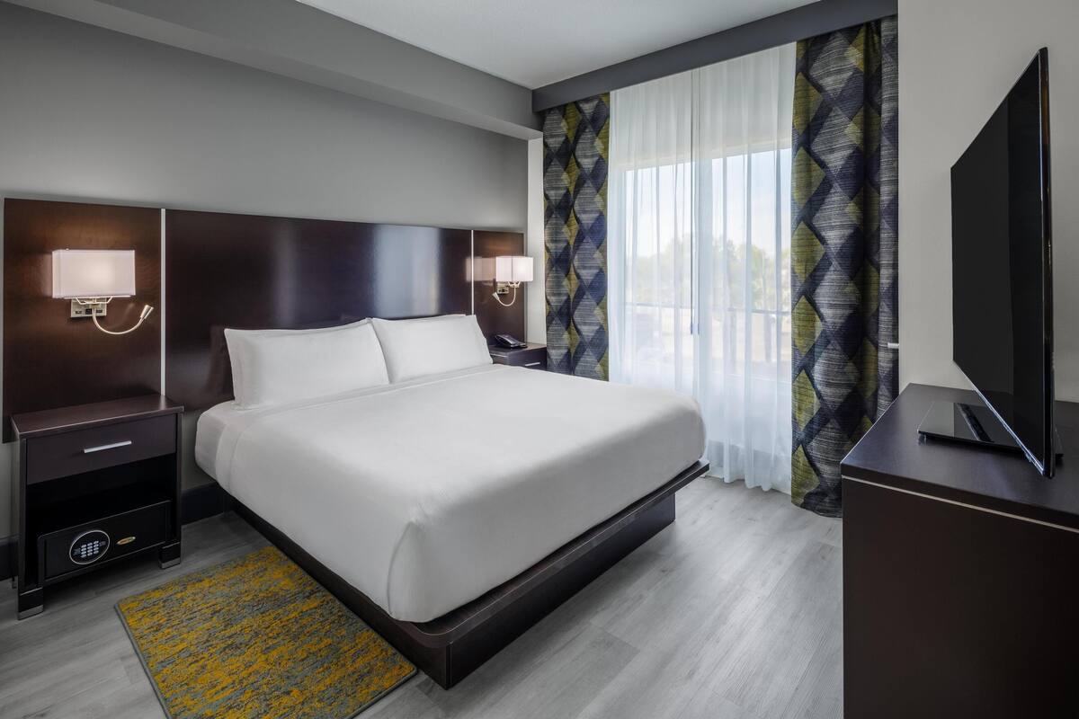 Een hotelkamer met een groot bed, nachtlampjes, een flatscreen-tv en een raam met gordijnen met een patroon.
