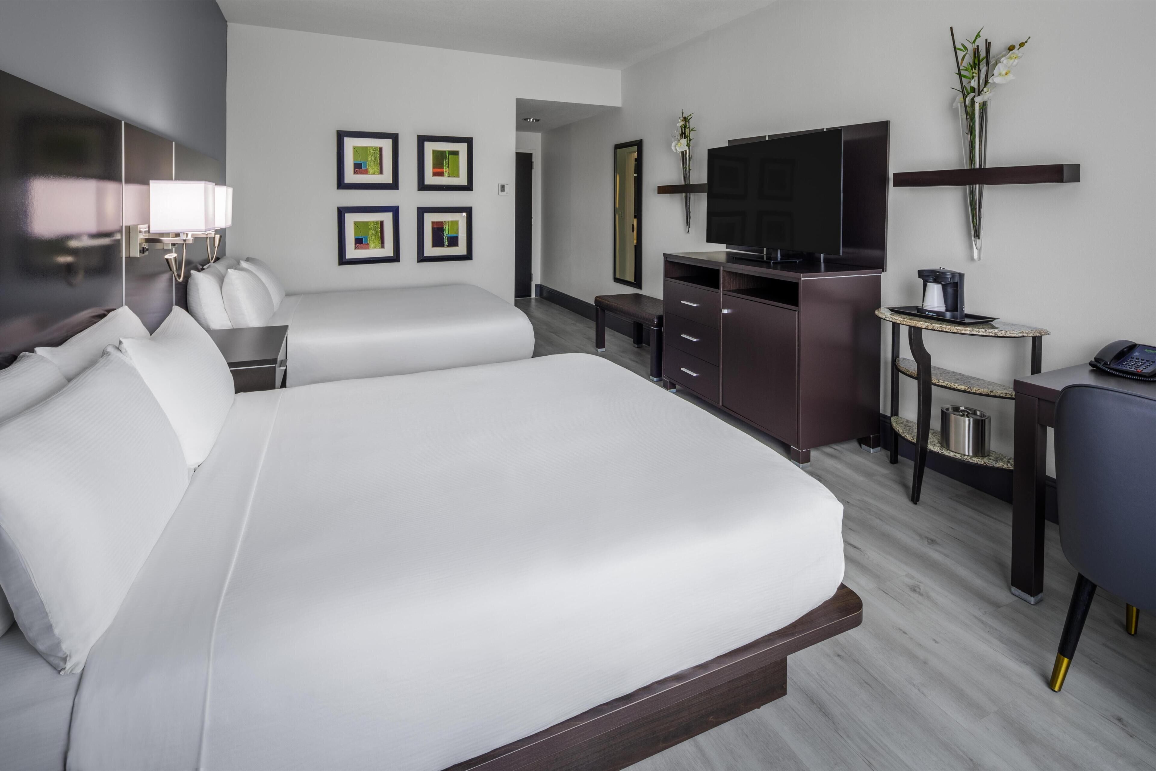 Une chambre d’hôtel avec deux lits, un bureau, une chaise, une télévision, un miroir et des œuvres d’art accrochées au mur.