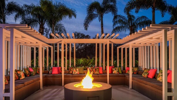 A circular fire pit surrounded by a wooden pergola with cushioned seating and colorful pillows.