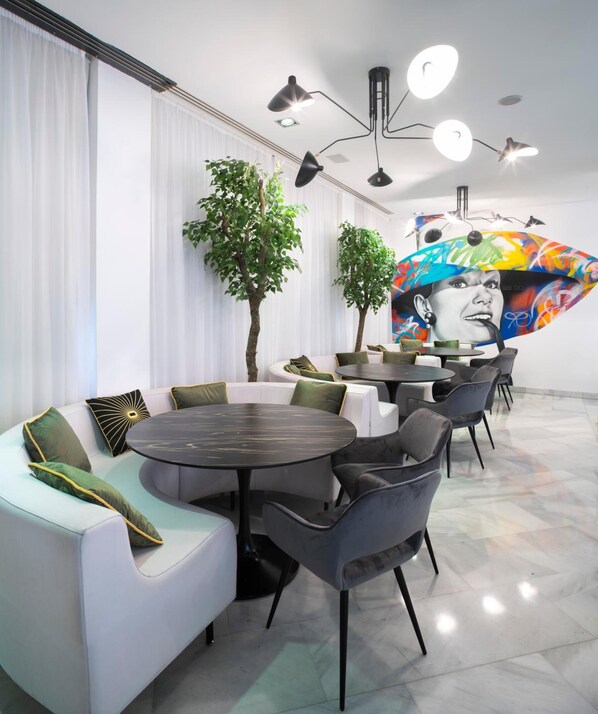 Un espace de détente moderne, aménagé avec des sièges, notamment des canapés et des fauteuils, et une table ronde. On y trouve deux plantes en pot et une fresque colorée sur le mur.