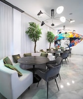 A modern lounge area with seating arrangements, including sofas and chairs, and a round table. There are two potted plants and a colorful mural on the wall.