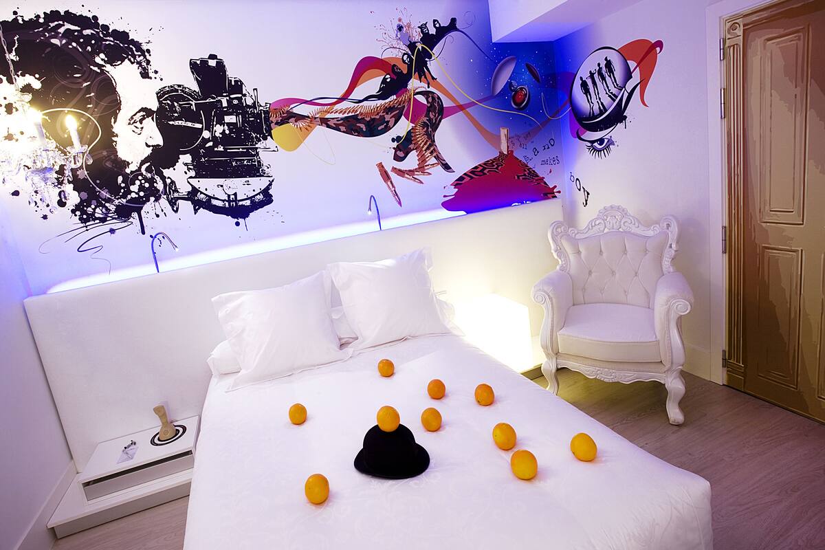 Ein Schlafzimmer mit einem Bett, weißen Kissen, einem weißen Stuhl und einer Wand mit abstrakter Kunst.
