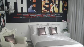 Une chambre avec un grand lit, un fauteuil blanc recouvert de tissu et un mur orné d’une grande affiche.