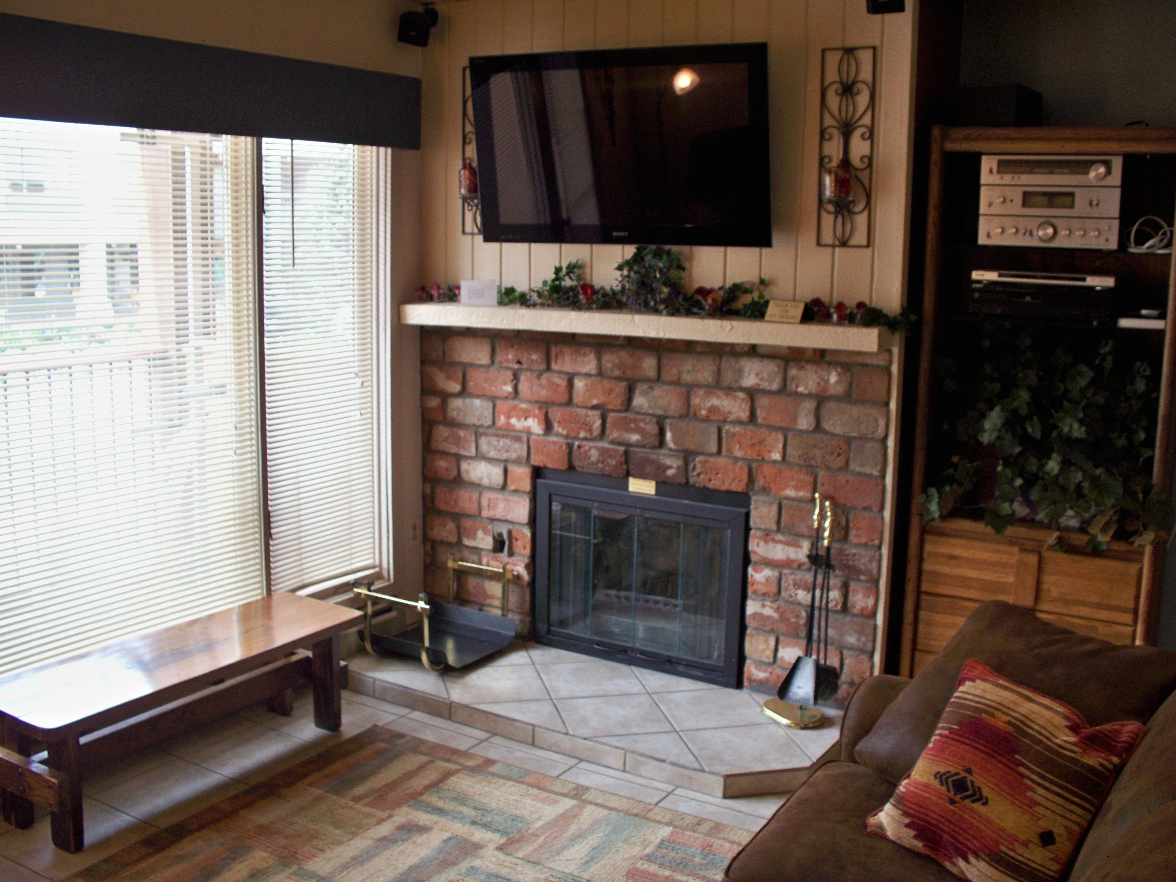 A living room with a brick fireplace, a flat-screen TV mounted above it, a wooden bench, a patterned rug, and a couch with a decorative pillow.