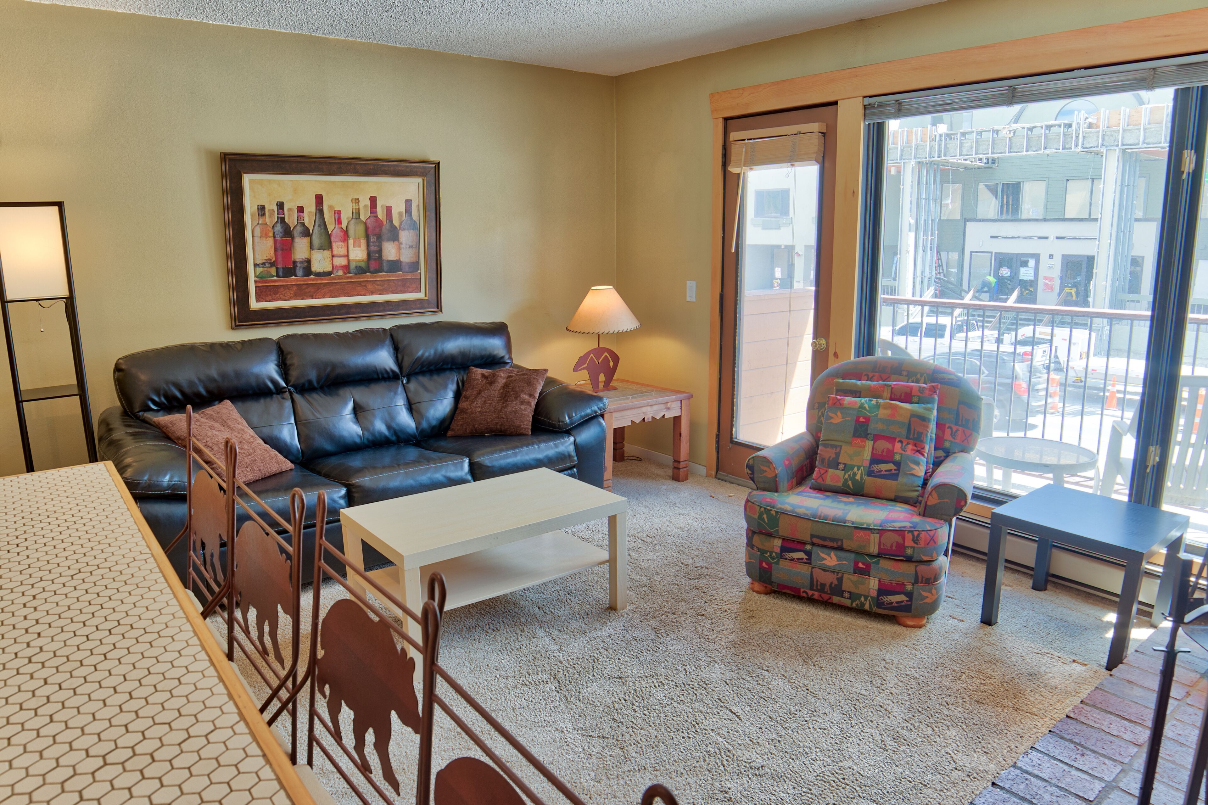 A living room with a black leather sofa, a colorful armchair, a white coffee table, and a painting of wine bottles on the wall.