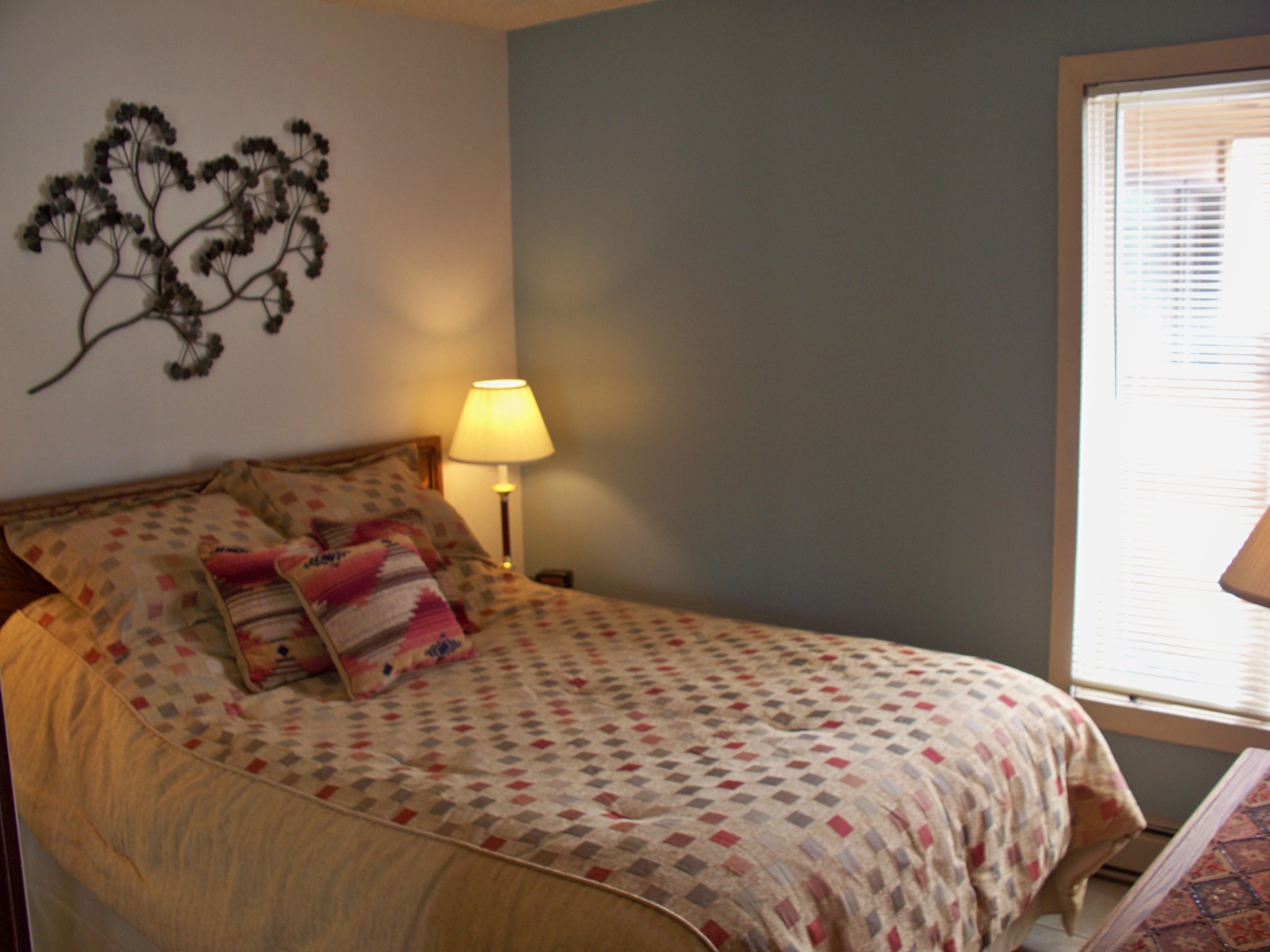 A bedroom with a bed, a wall-mounted plant, a bedside lamp, and a window with blinds.