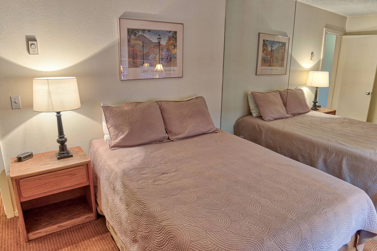 Une chambre d’hôtel avec deux lits, chacun doté d’une table de chevet et d’une lampe, un tableau encadré au mur et une petite étagère sous le lit situé à gauche.