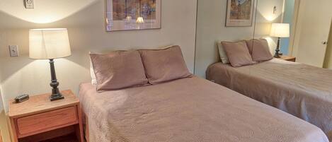 A hotel room with two beds, each with a bedside table and lamp, a framed picture on the wall, and a small shelf under the left bed.