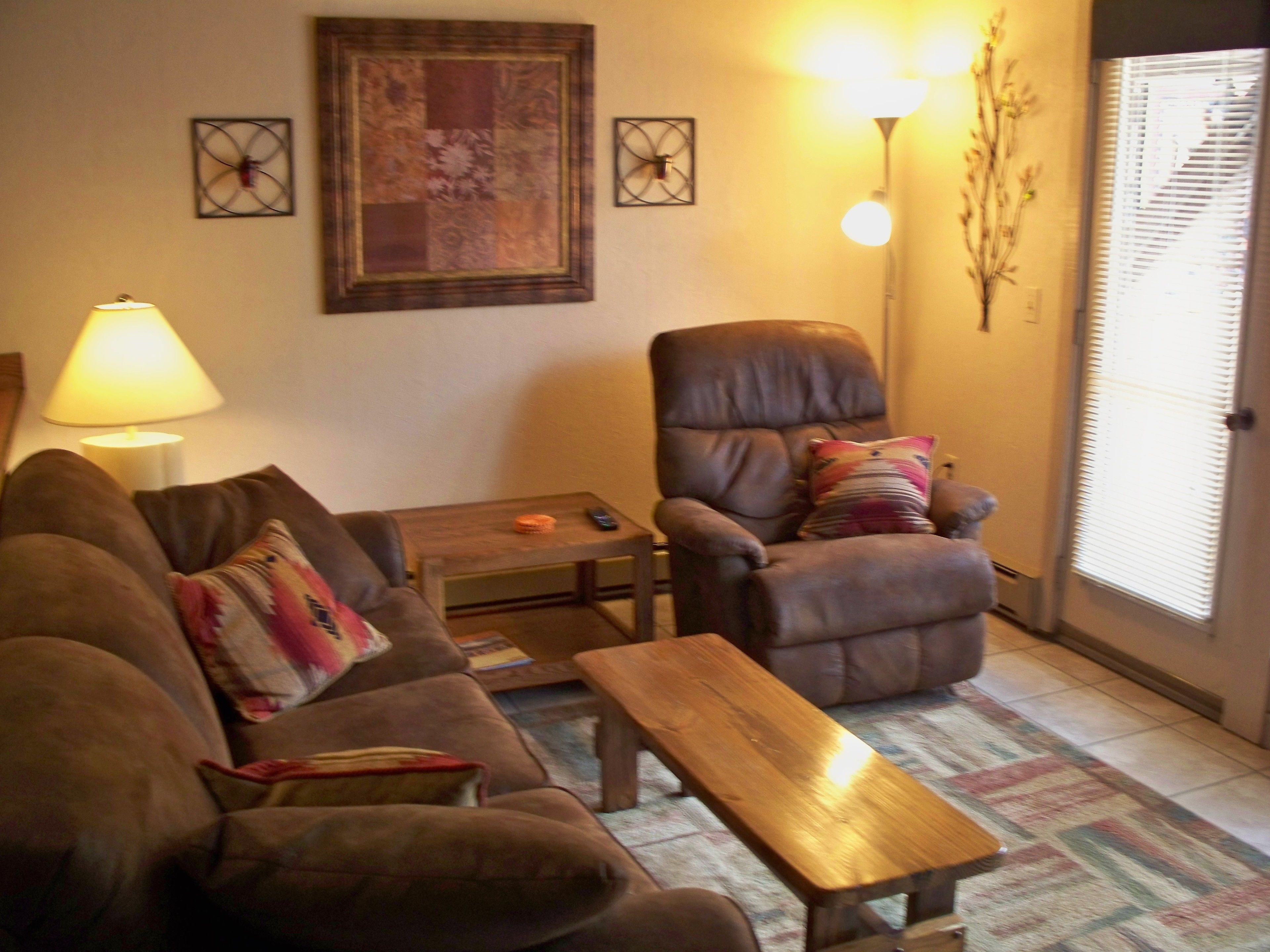 Ein Wohnzimmer mit einem braunen Ledersofa, einem Holztisch, einem braunen Sessel mit einem farbenfrohen Kissen und einer Lampe.