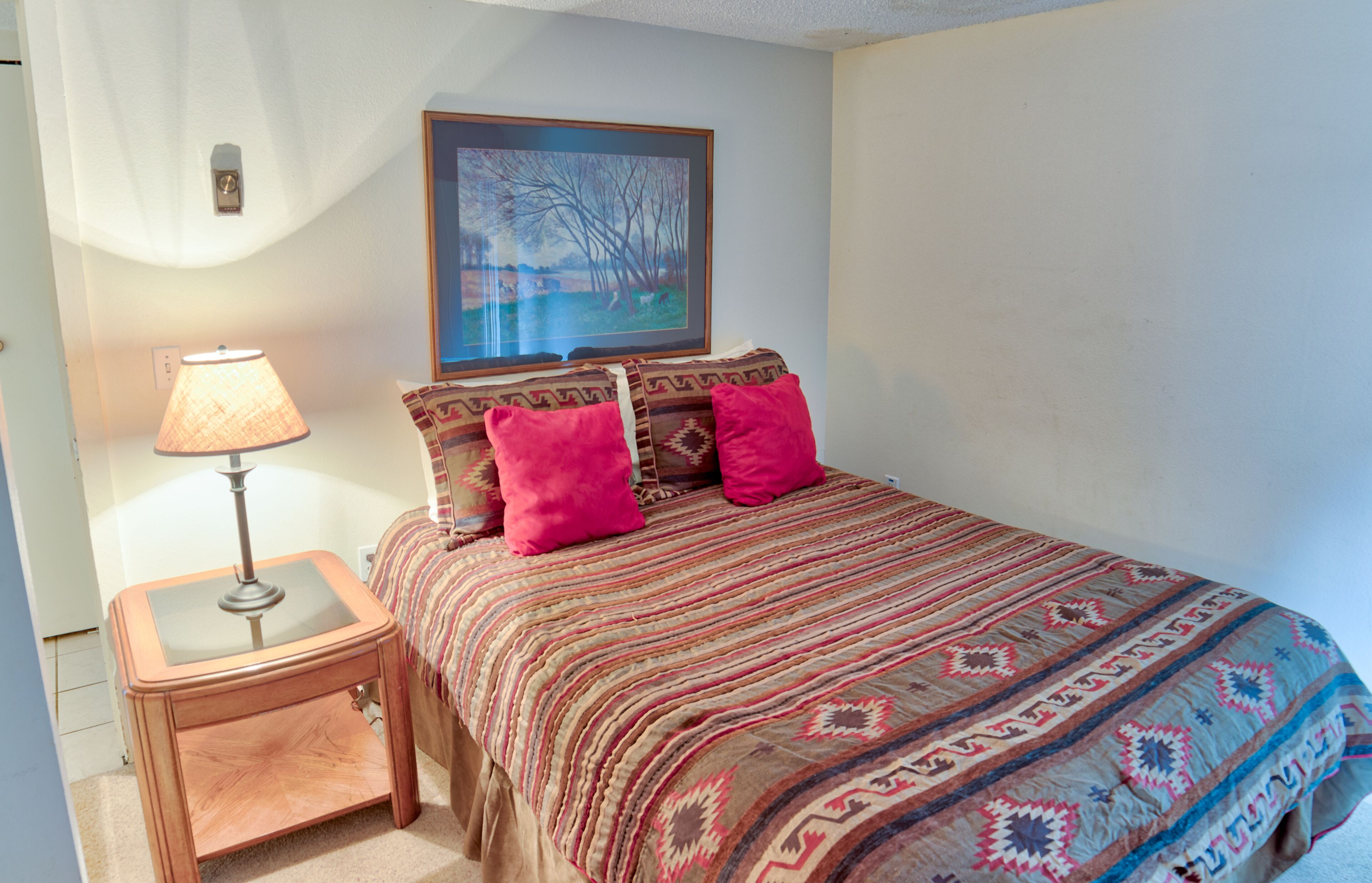 Un dormitorio con cama, mesita de noche, lámpara y un cuadro en la pared.