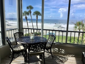 Ein Balkon mit rundem Tisch und Stühlen, der Blick auf einen Strand mit Palmen und einem klaren Himmel gewährt.