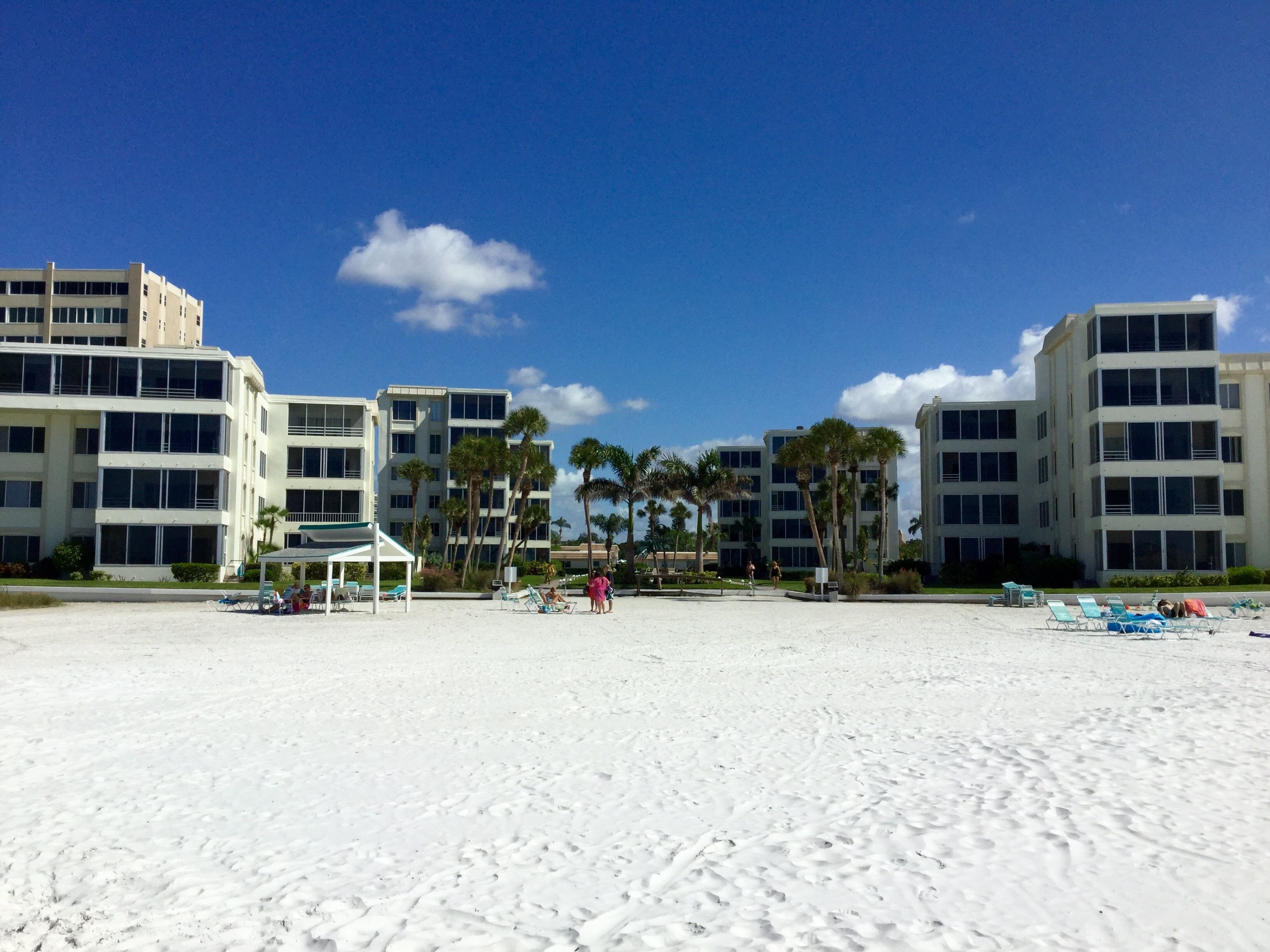 A beach with white sand, palm trees, and a building with multiple floors and balconies.