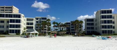 A beach with white sand, palm trees, and a building with multiple floors and balconies.