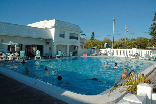 A swimming pool area with lounge chairs and umbrellas, a white building with multiple windows, and an American flag.