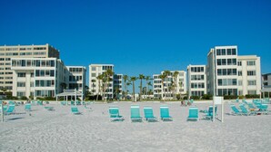 Un hotel frente a la playa con varios pisos, una playa de arena con sillas de descanso azules y palmeras.