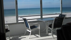 Una habitación con un ventanal grande con vista a la playa. Hay una mesa y dos sillas con cojines azules.