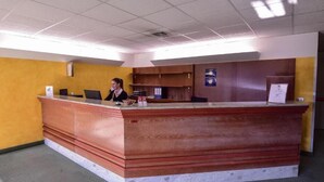 A reception desk with a receptionist, a wooden counter, and a yellow wall. - Standard Hotel Udine (Pradamano)