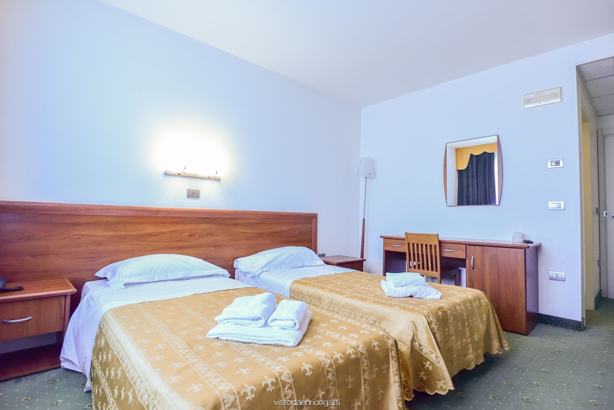 Camera d'albergo con due letti, una scrivania, una sedia e uno specchio.