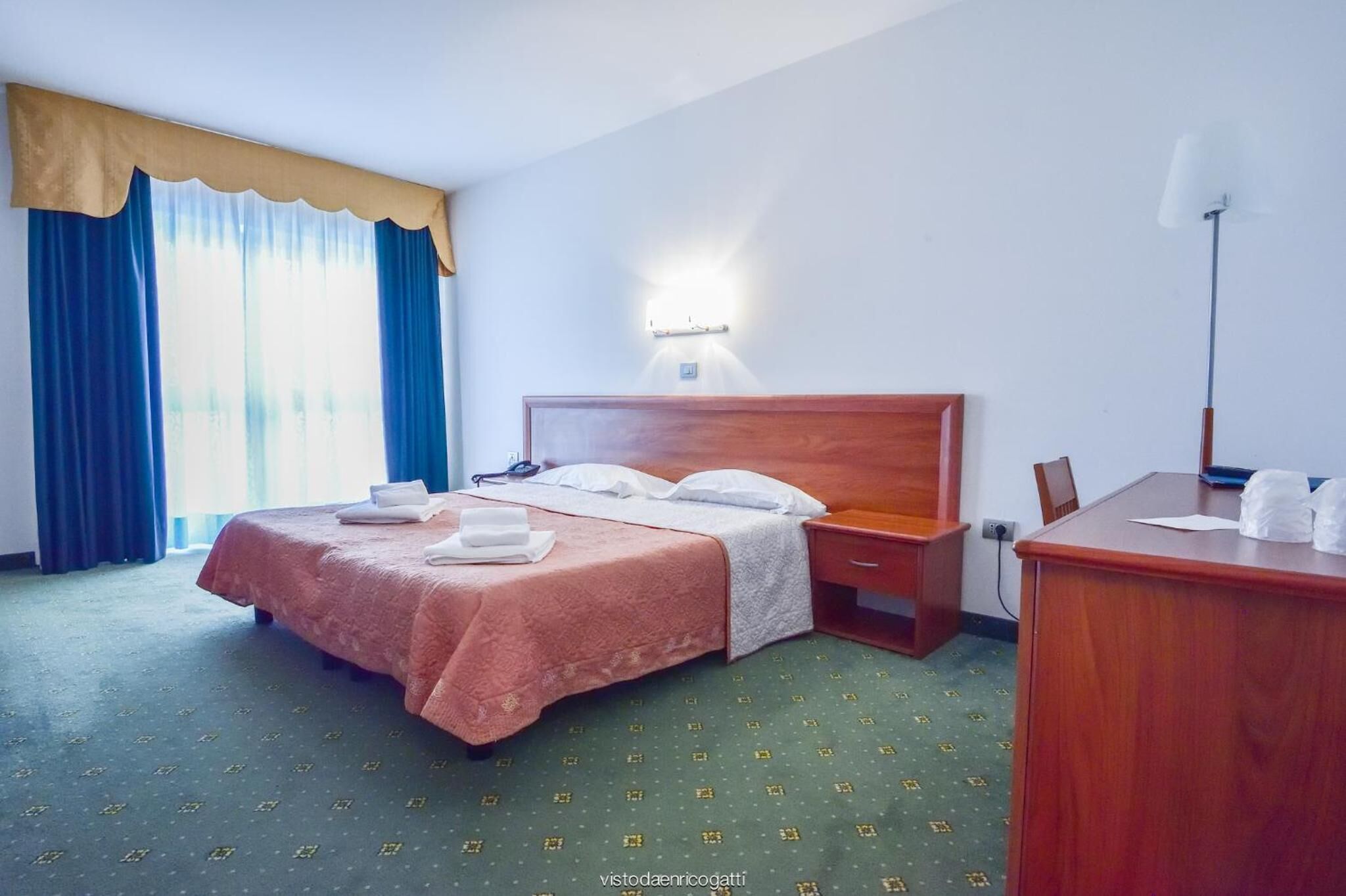 A hotel room with a double bed, a bedside table, a desk with a lamp, and a large window with curtains.