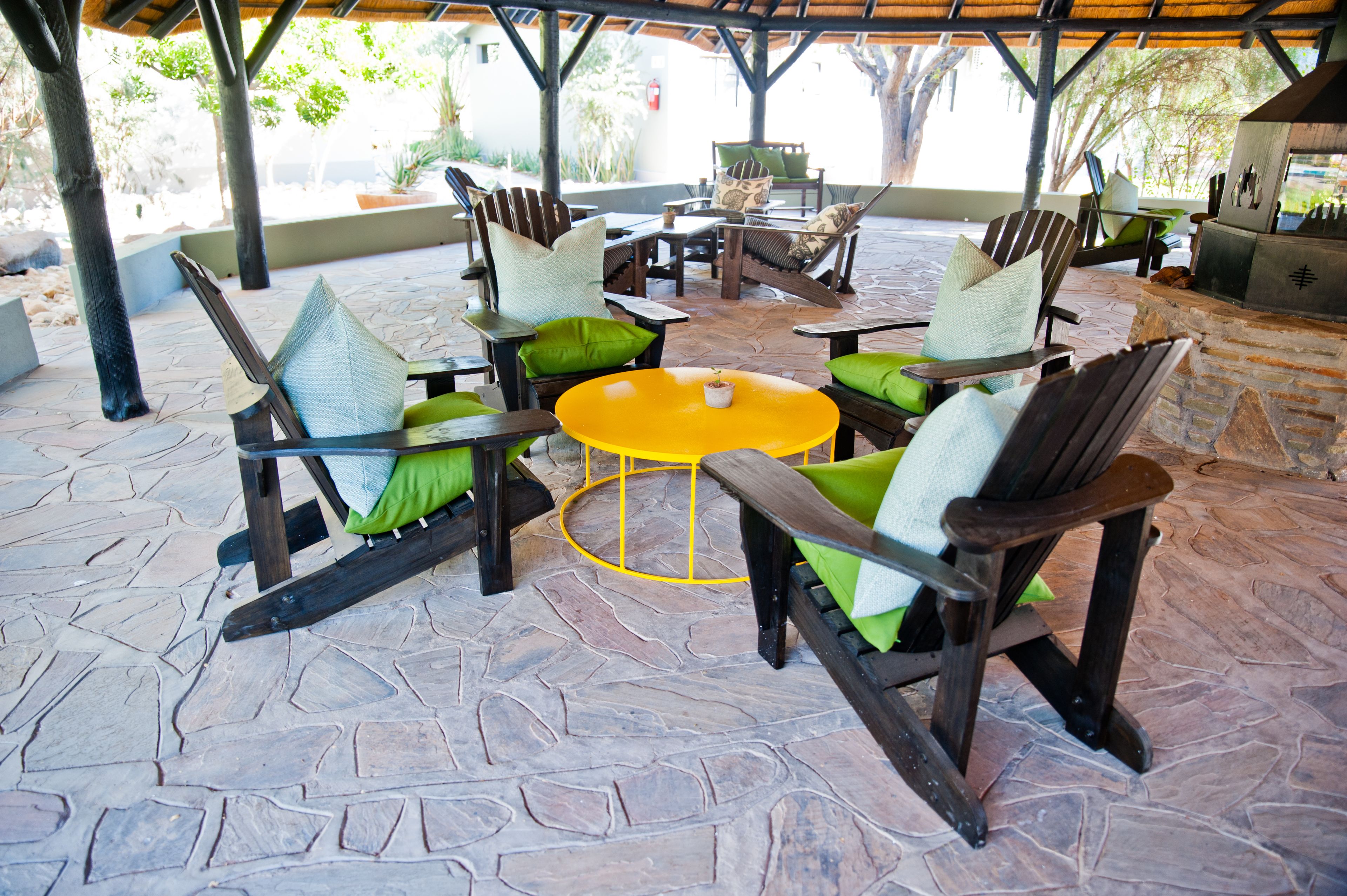 An outdoor patio with wooden chairs and a yellow table, surrounded by stone flooring.