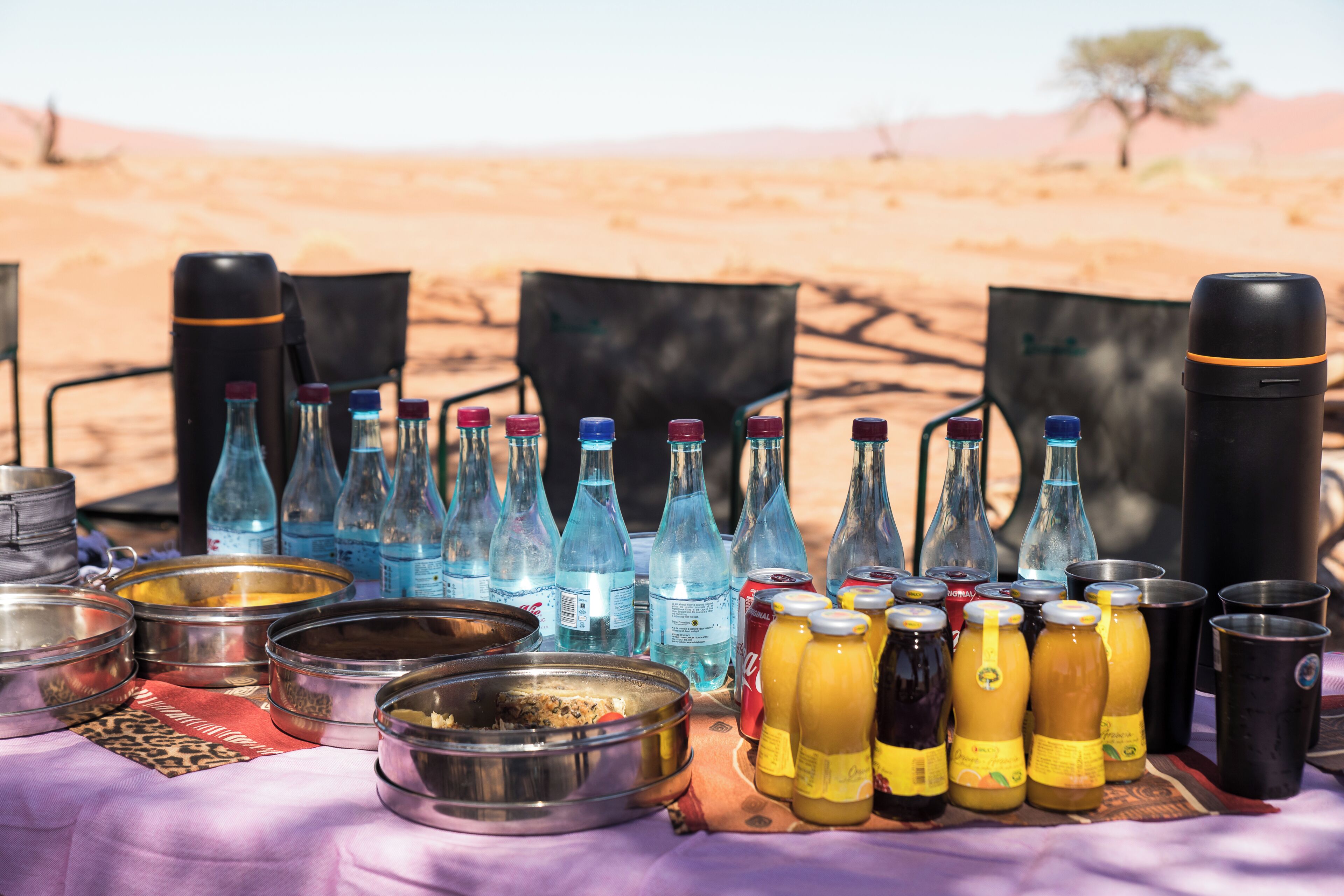 A desert campsite with a table set up with various food containers, bottles, and a thermos.