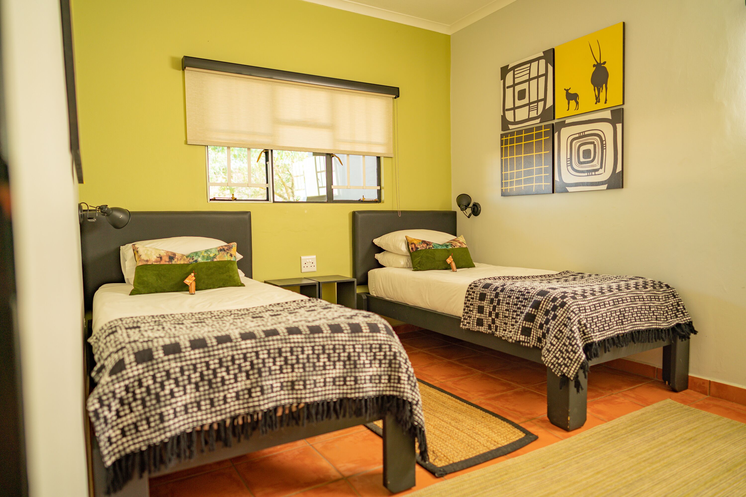 A room with two beds, each with a patterned bedspread and pillows, a window with a blind, and artwork on the wall.