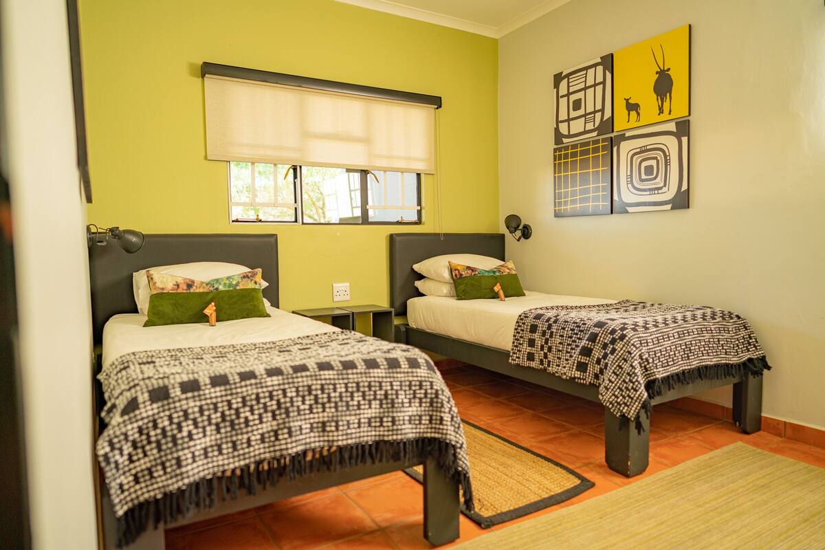 A room with two beds, each with a patterned bedspread and pillows, a window with a blind, and artwork on the wall.