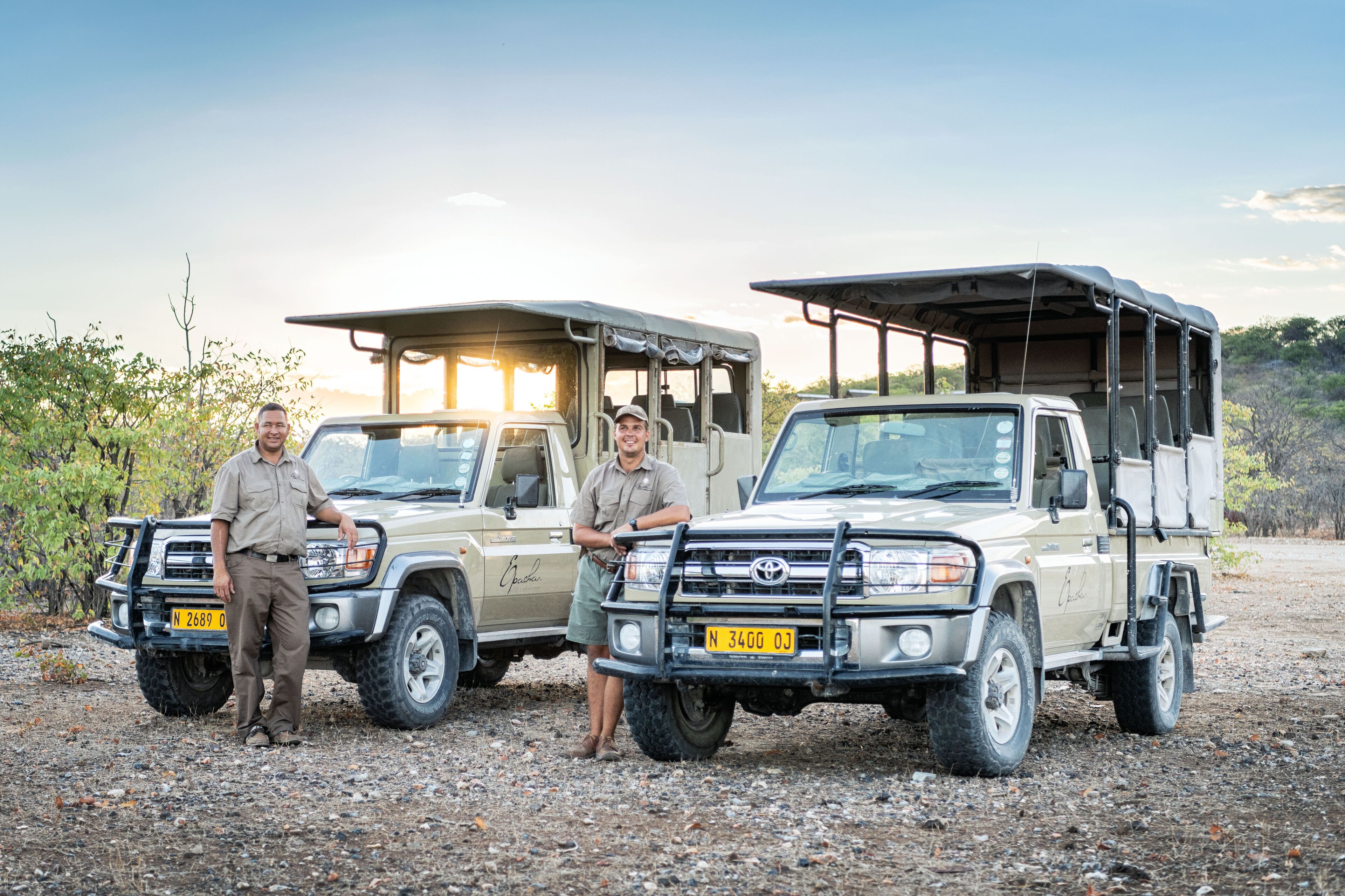 Two safari vehicles with a rugged, off-road design, equipped with roof racks and side storage compartments, parked on a gravel surface with sparse vegetation in the background.