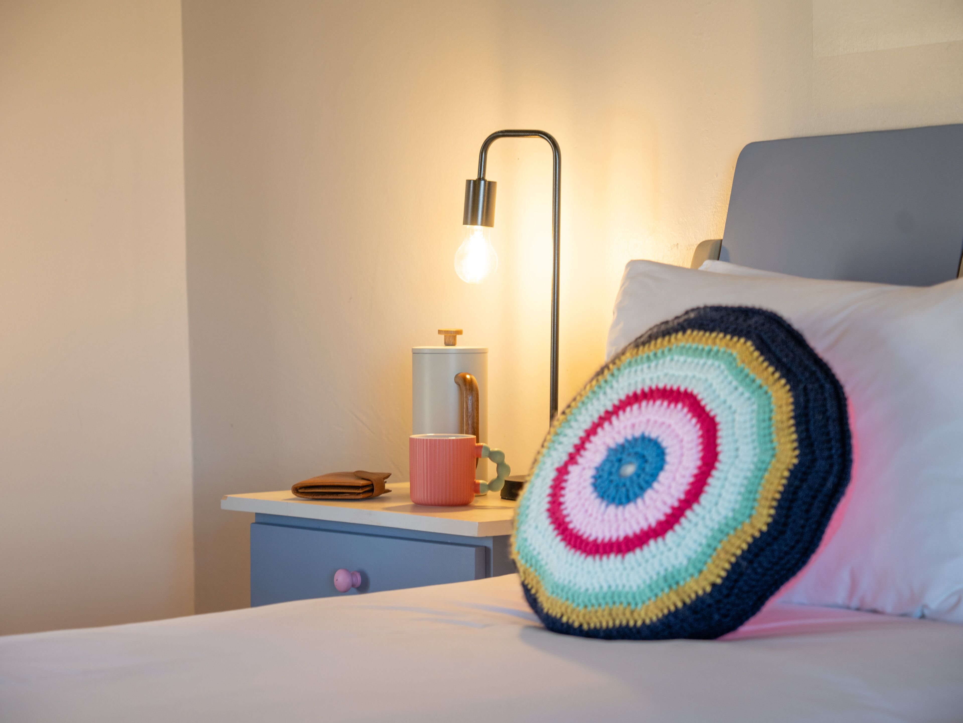 A neatly made bed with a colorful crocheted pillow, a bedside table with a lamp, and a cup.