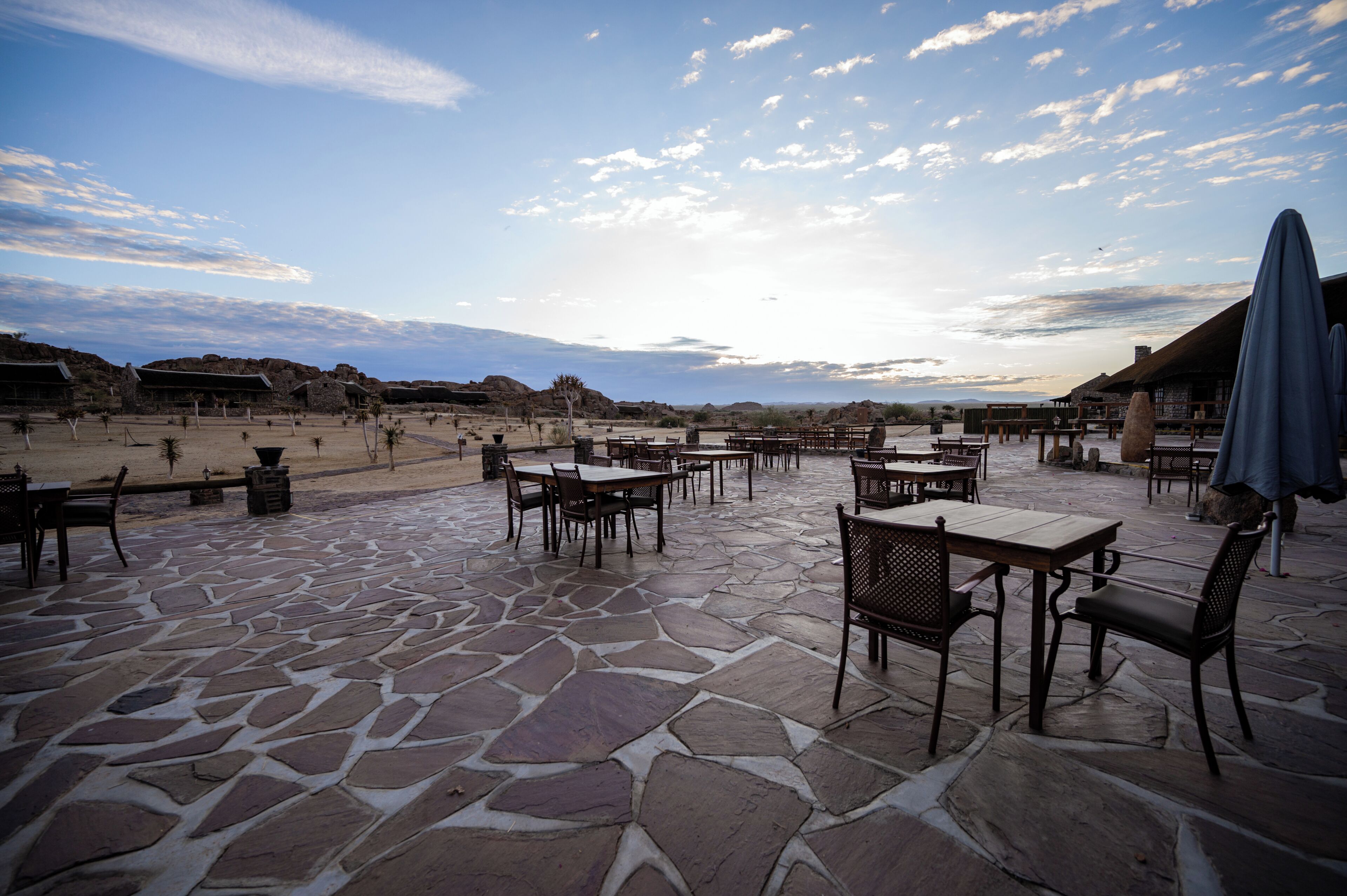 Un patio al aire libre con suelo de piedra, mesas y sillas, con vistas a un paisaje desértico.
