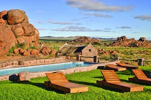 Zona de piscina con sillones de descanso, un edificio de piedra y un paisaje rocoso al fondo.