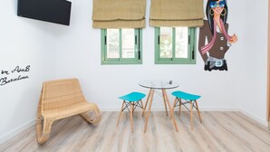 A room with a wicker chair, a small round table with two blue chairs, two windows with beige curtains, and a wall-mounted TV. - AinB Las Ramblas Colón Apartments (Barcelona)
