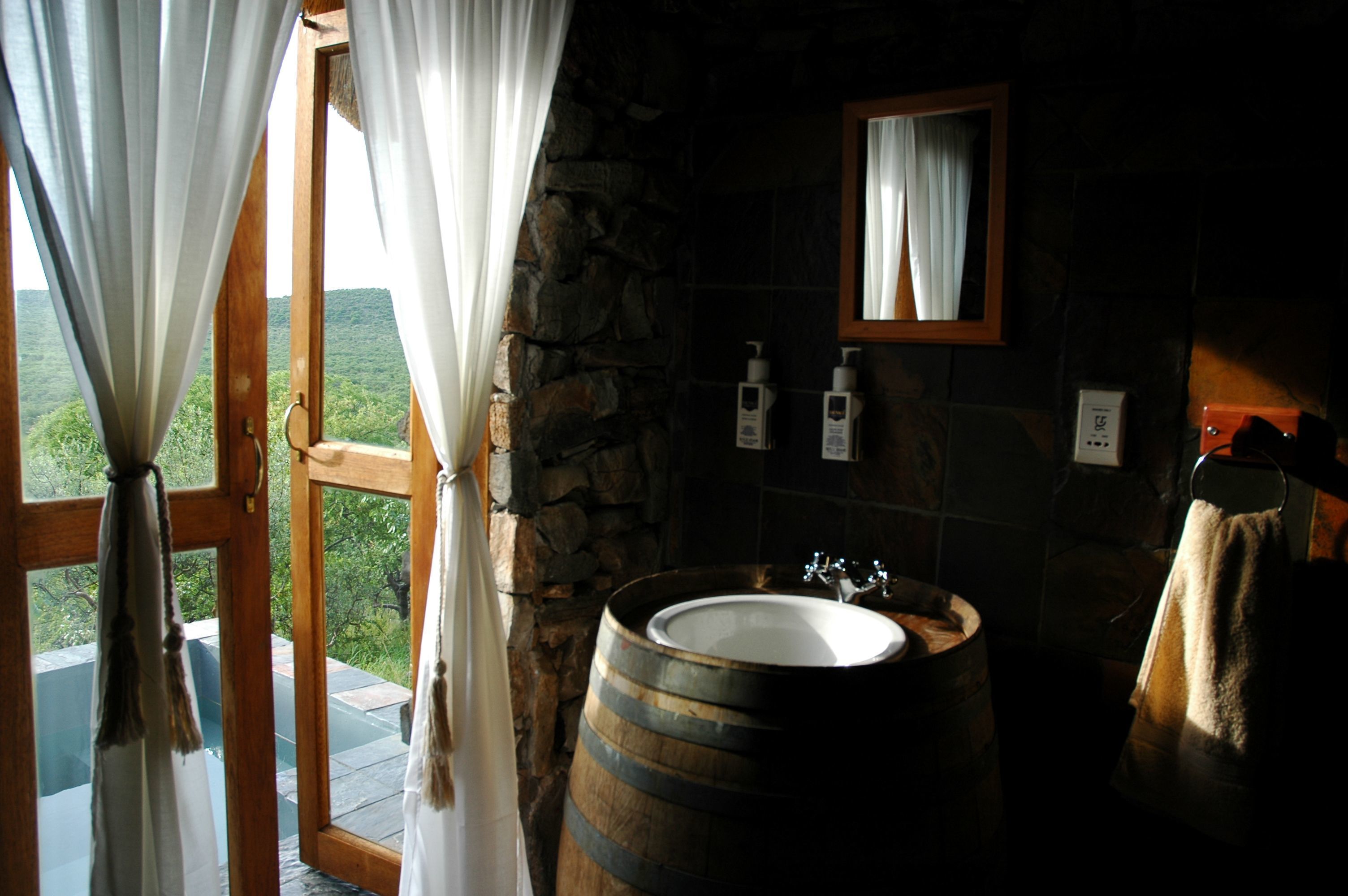 A bathroom with a stone wall, wooden barrel sink, and a view through a window with curtains.