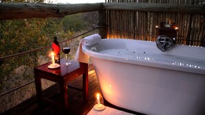 Eine Holzterrasse mit einer weißen Badewanne, einem kleinen Tisch mit Kerze und einem Glas Rotwein.