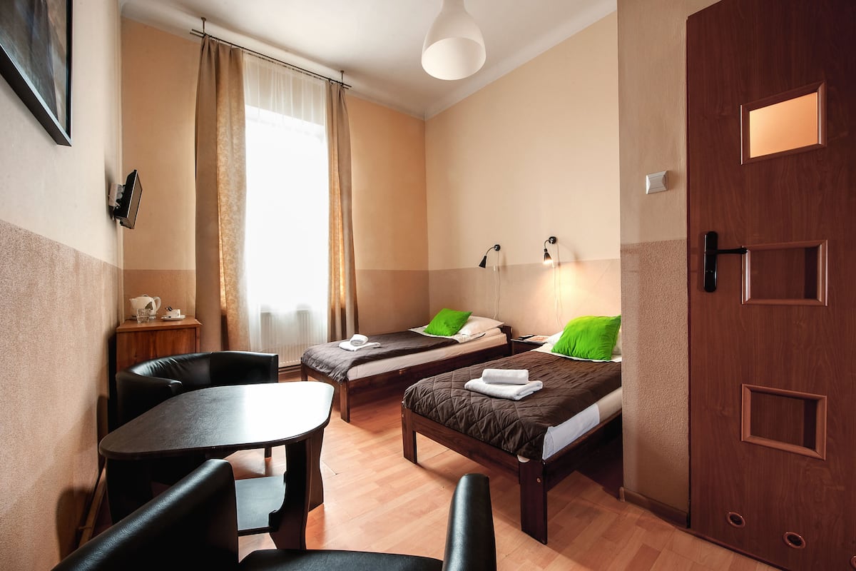 Une chambre d’hôtel avec deux lits simples, une table en bois, une chaise, une petite table de chevet et une fenêtre avec des rideaux.