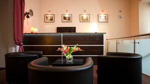 Un vestíbulo de hotel con un mostrador de recepción de madera oscura, dos sillones negros, un jarrón de flores rojas y cuadros enmarcados en la pared.