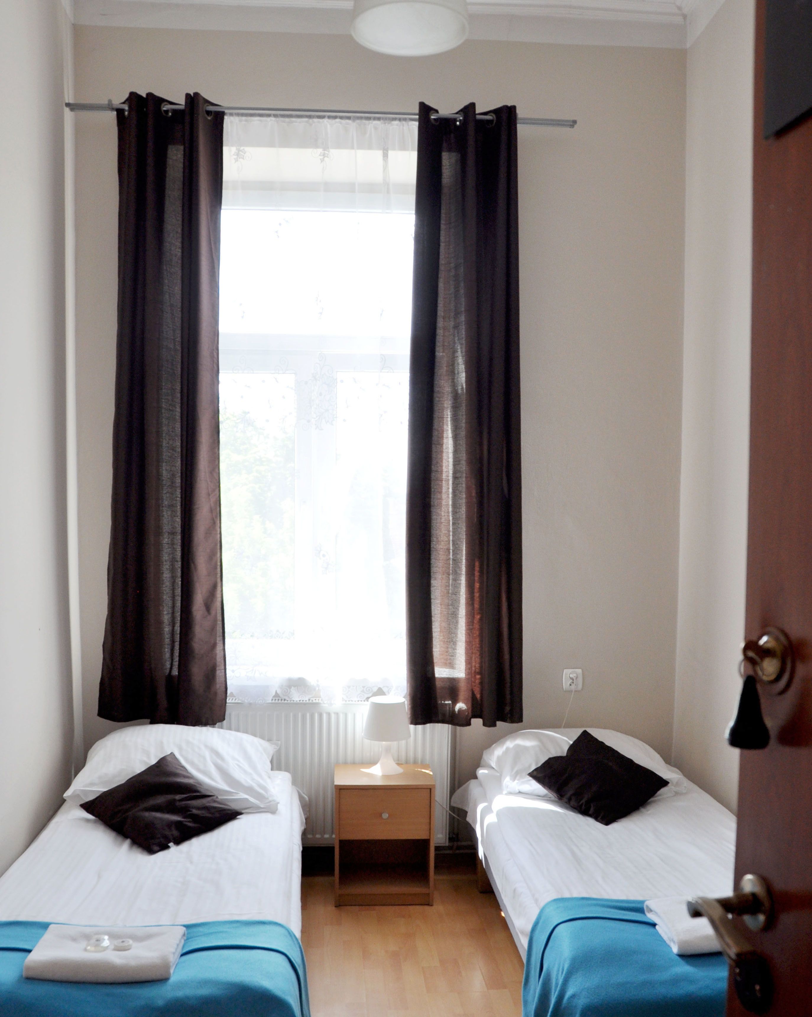 A small, single-room hotel room with two single beds, a nightstand with a lamp, and a window with curtains.